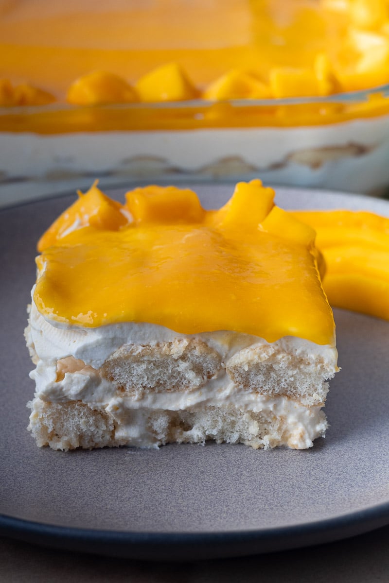 Slice of creamy mango tiramisu on a gray plate featuring layers of cream and sponge topped with fresh mango slices.