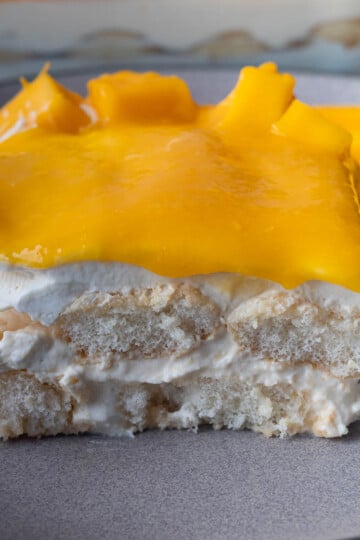 Slice of creamy mango tiramisu with cream, sponge cake, and bright yellow mango topping on a plate.