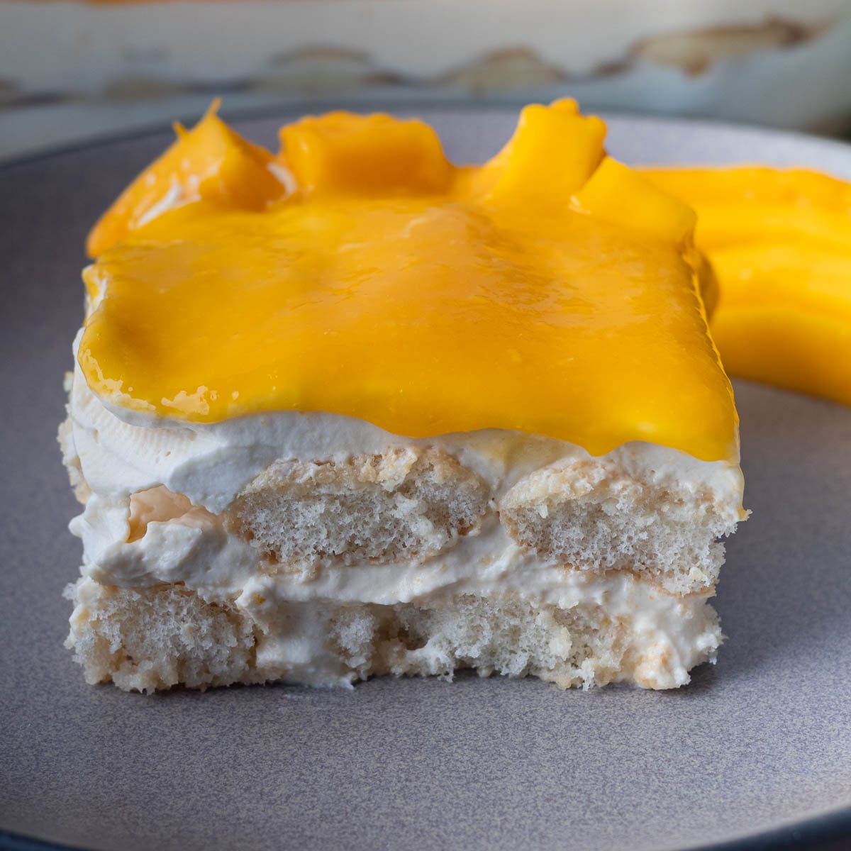 Slice of creamy mango tiramisu with cream, sponge cake, and bright yellow mango topping on a plate.