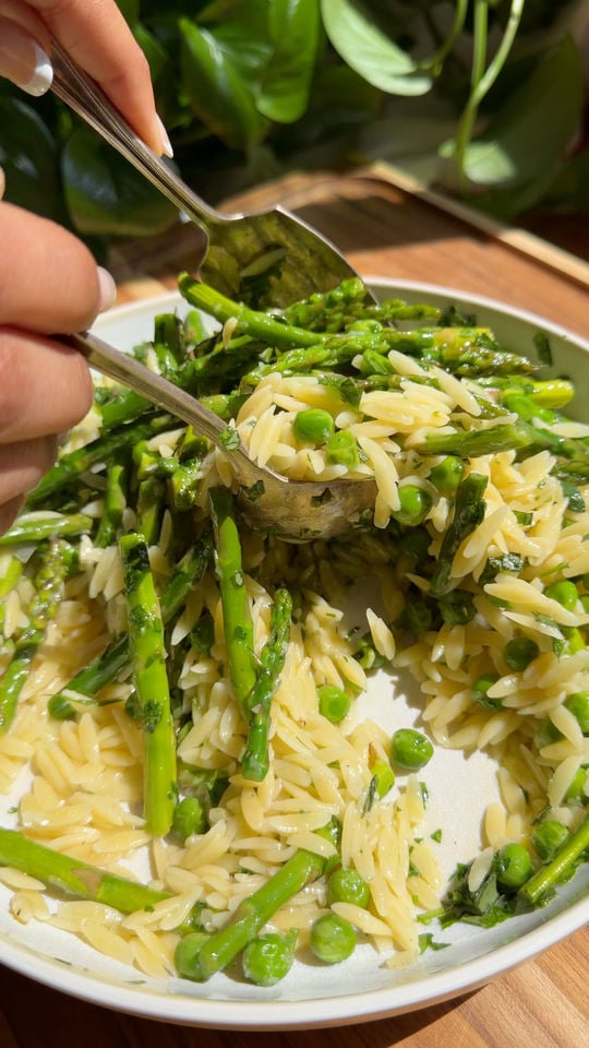 Orzo pasta being tossed with asparagus and peas in a bowl.