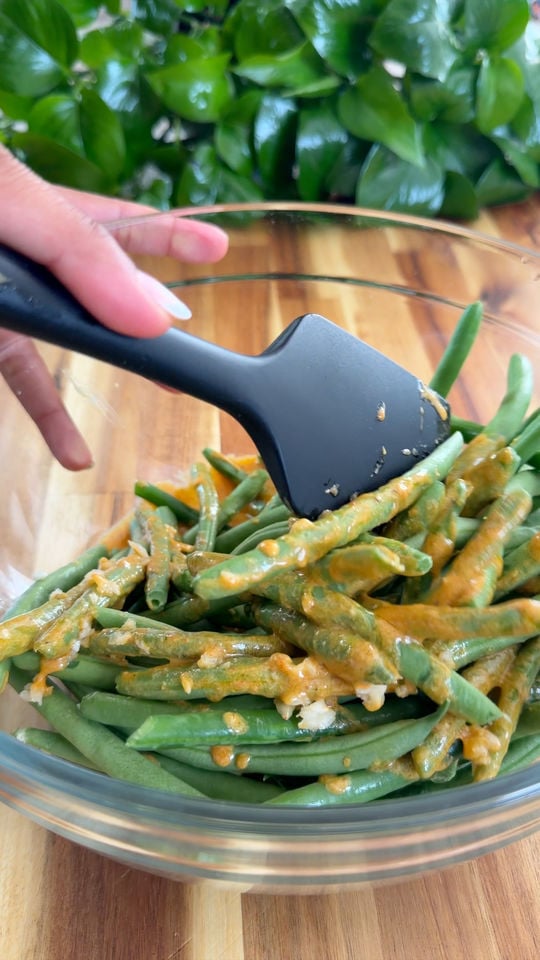 Mixing fresh green beans with sauce in a glass bowl using a black spatula.