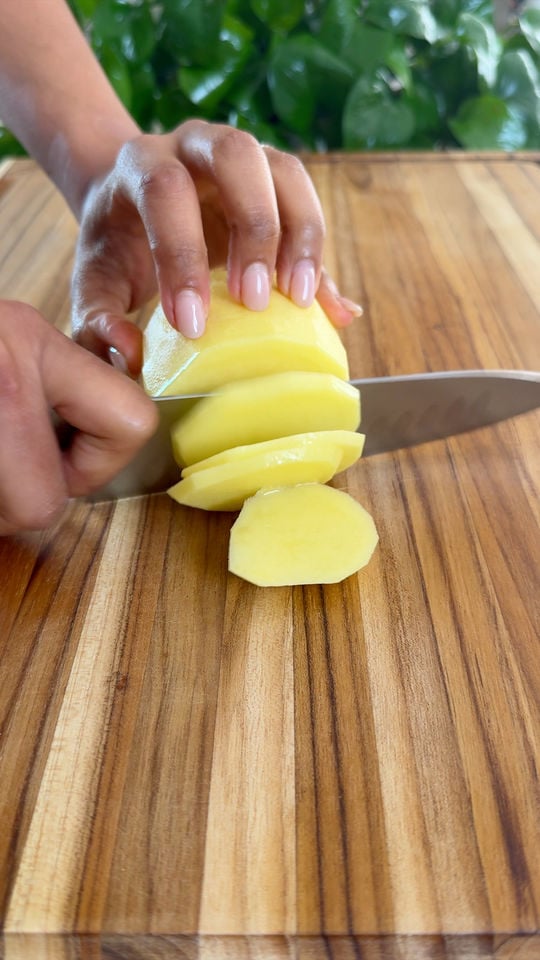 Slicing a peeled potato with a knife on a wooden cutting board.