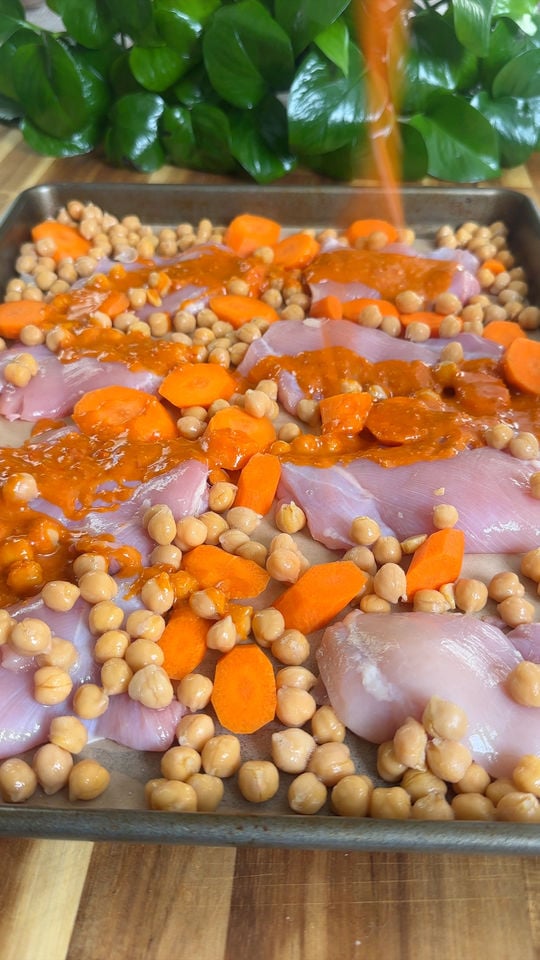Raw chicken, carrots, and chickpeas on a baking tray being drizzled with a harissa sauce.
