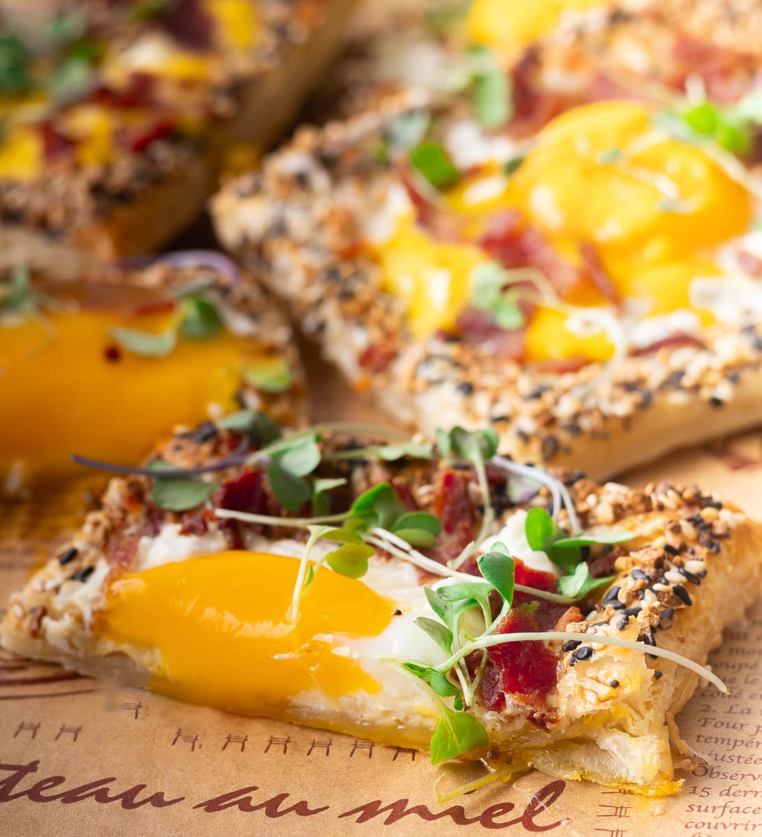 Close-up of a puff pastry breakfast pizza with egg yolk, bacon, sesame seeds, and fresh microgreens on top.