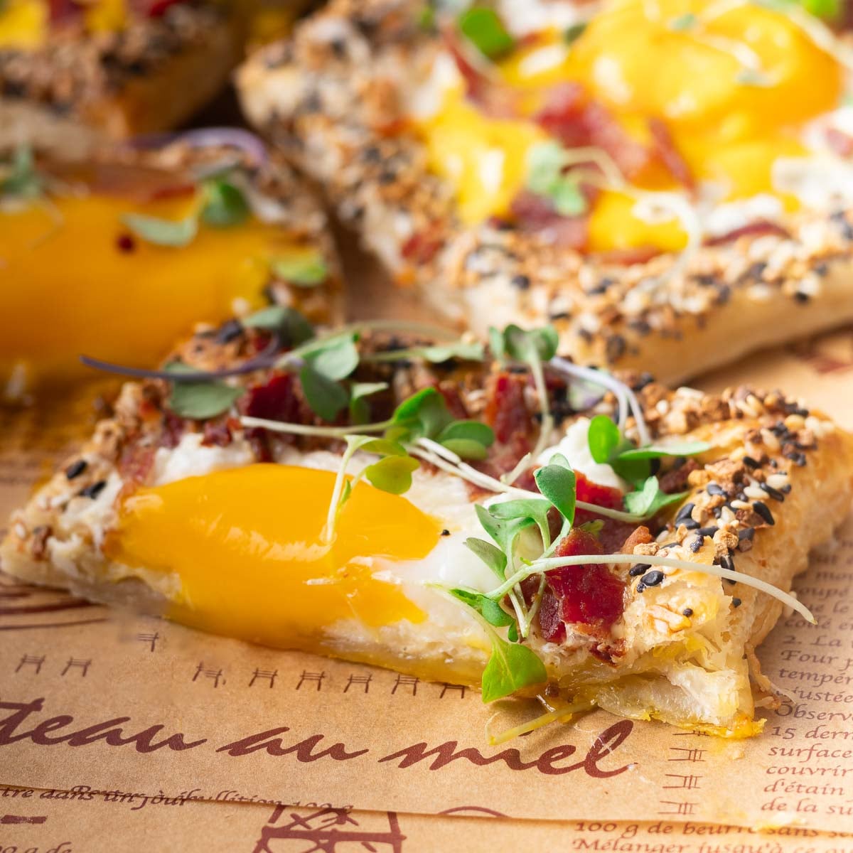 Close-up of a puff pastry breakfast pizza sesame seeds, melted cheese, runny egg yolk, and microgreens on a parchment paper.