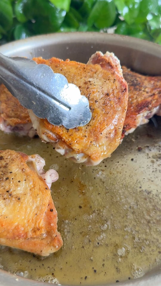Tongs holding a seasoned, golden-brown chicken thigh in a pan with oil.