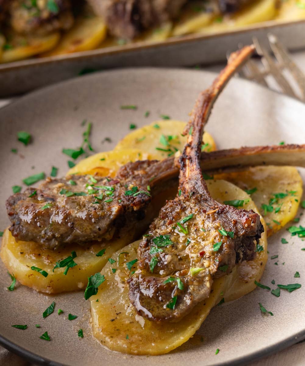 Sheet pan baked lamb chops on sliced potatoes, garnished with chopped parsley, served on a gray plate.
