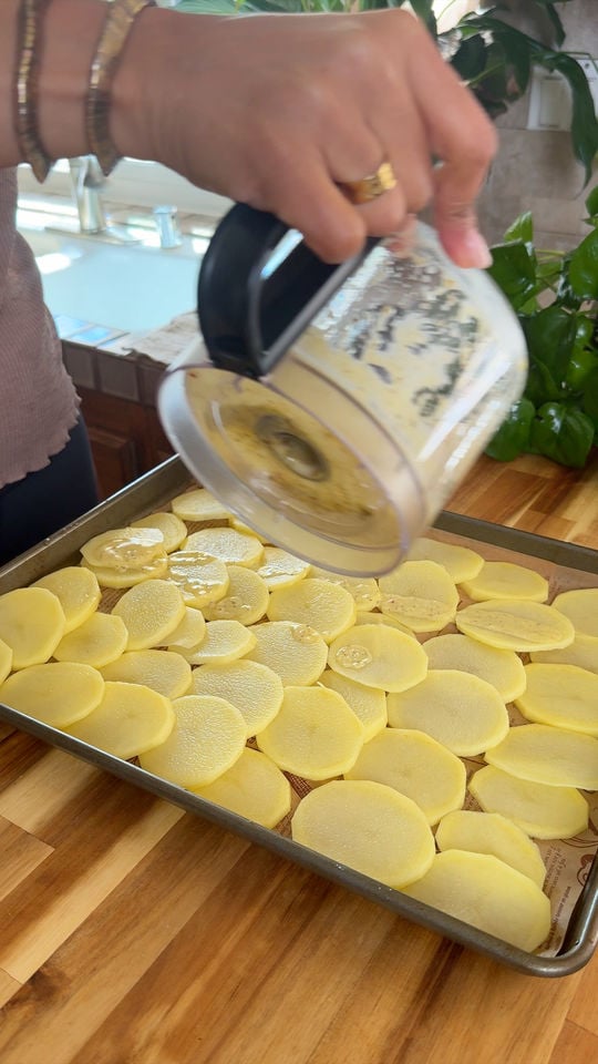 Hand pouring creamy sauce over sliced potatoes on a baking sheet.