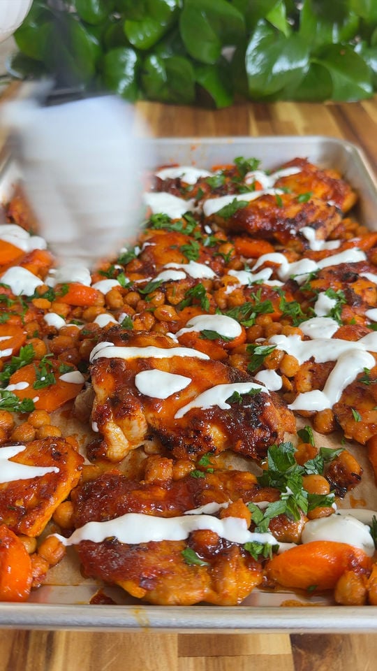 Baked chicken with chickpeas and carrots, topped with yogurt drizzle and fresh herbs on a tray.