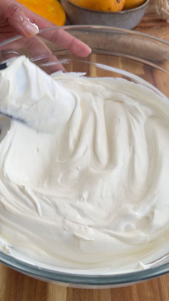 Hand stirring cream in a glass bowl on a wooden table.