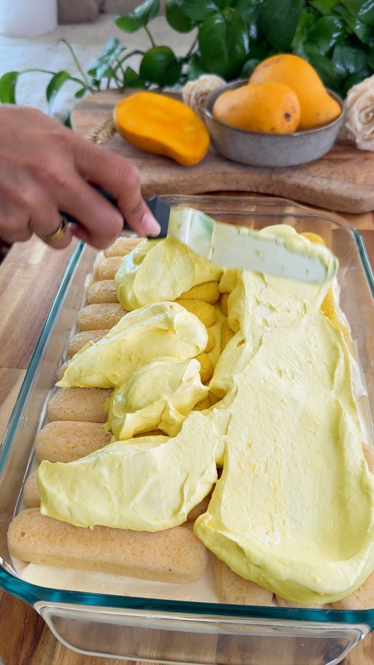 Hand spreading mango cream over ladyfingers in a glass dish.
