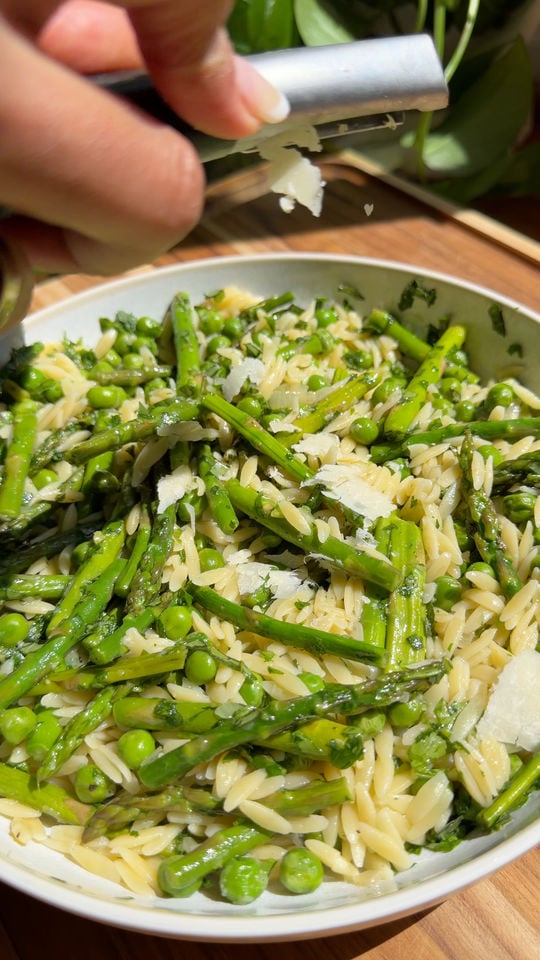Grating cheese over orzo with asparagus, peas, and herbs in a white bowl on a wooden table.
