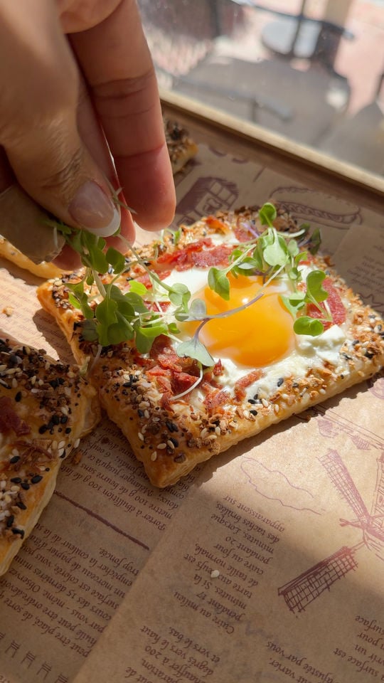 Hand garnishing pastry with egg, bacon, and microgreens on a wooden board.