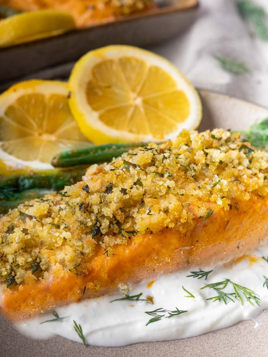 Baked panko crusted salmon filet on creamy sauce with dill, lemon slices, and green beans, served on a plate.