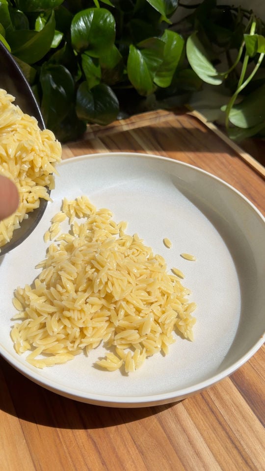 Orzo pasta being poured into a white bowl on a wooden table.
