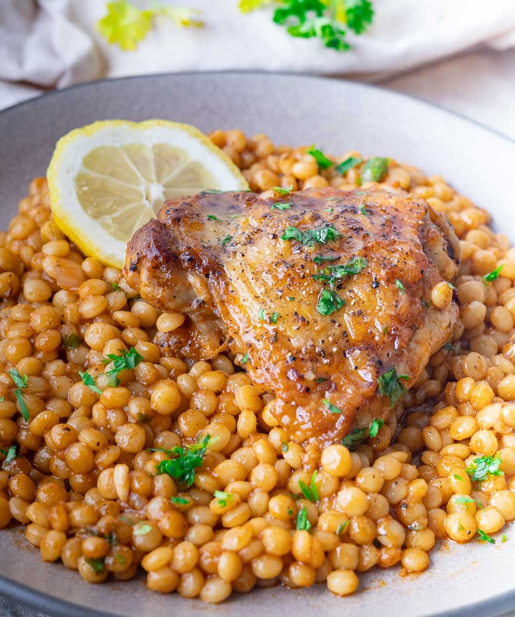 Roasted chicken thigh on a bed of spiced pearled couscous with parsley and lemon slice garnish.