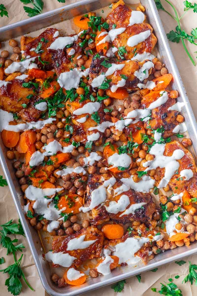 Honey harissa chicken tray bake with roasted chicken thighs with carrots, chickpeas, and drizzled sauce on a baking tray, garnished with parsley.