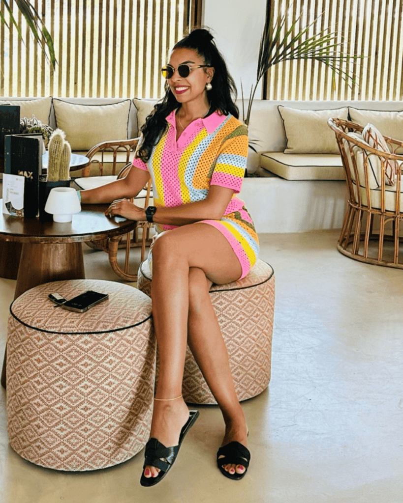 Woman in colorful dress sitting in a stylish lounge, smiling, with sunglasses and a smartphone nearby.