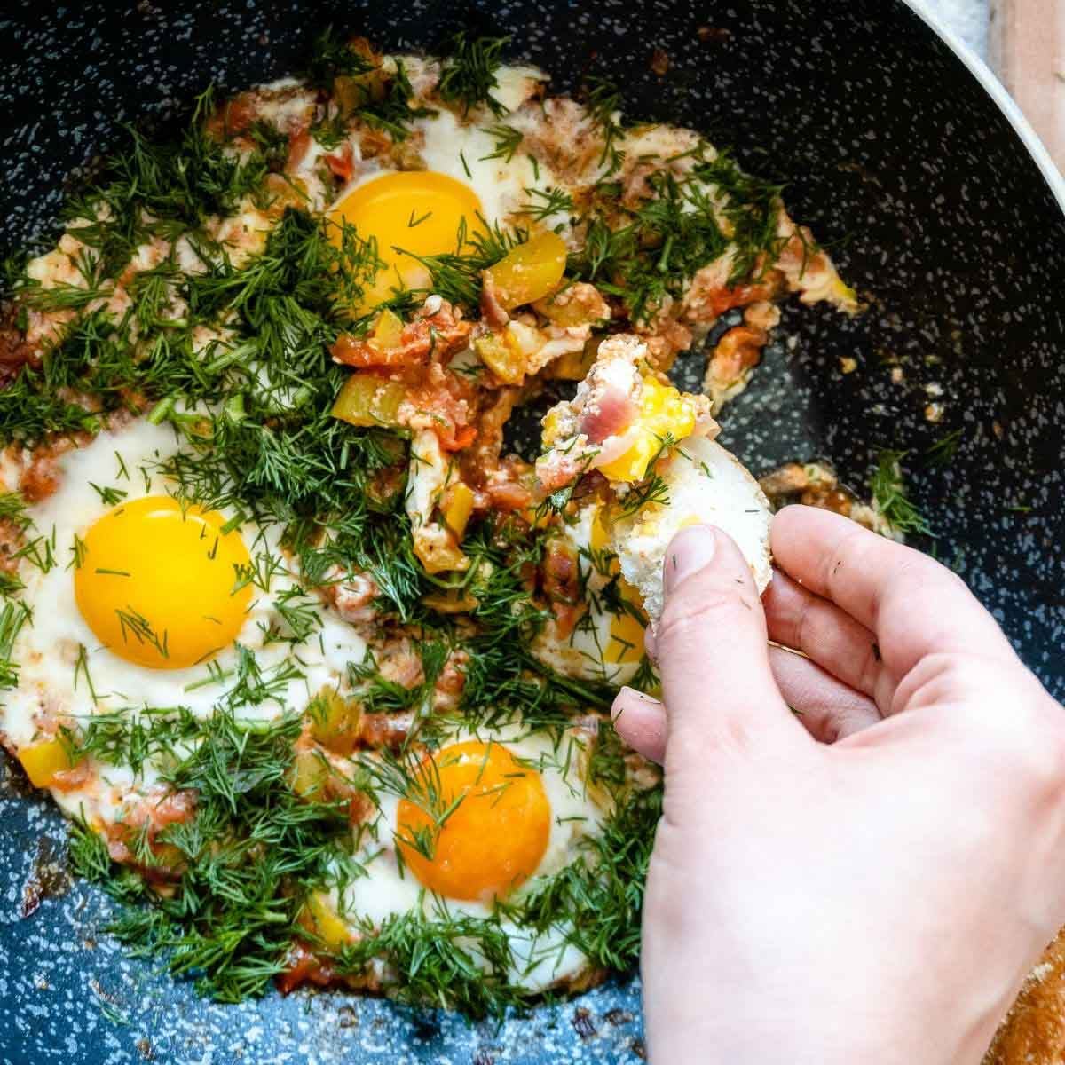Person dipping bread into a skillet with fried eggs, vegetables, and fresh herbs.
