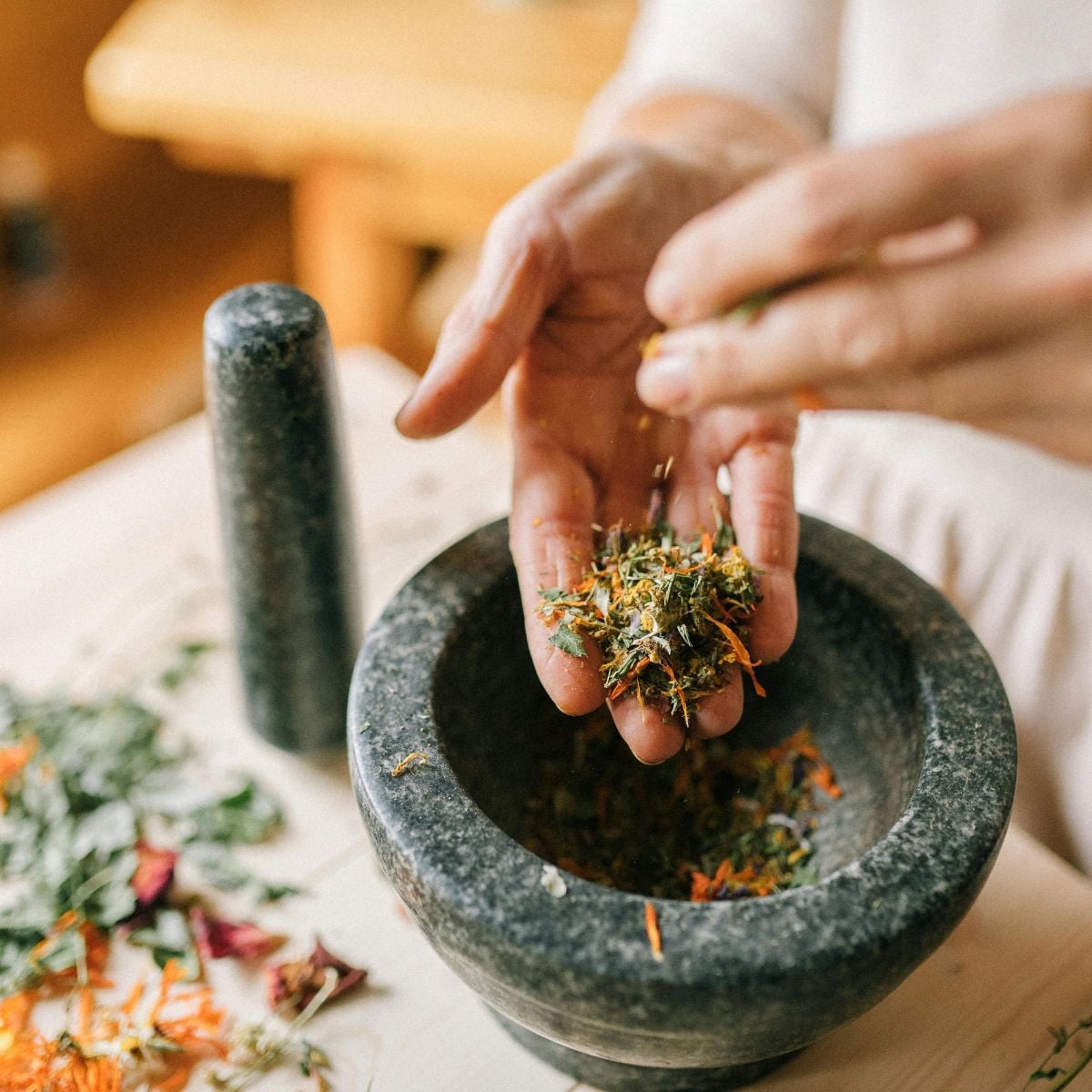 Hands holding dried herbs over a granite mortar and pestle on a wooden table.