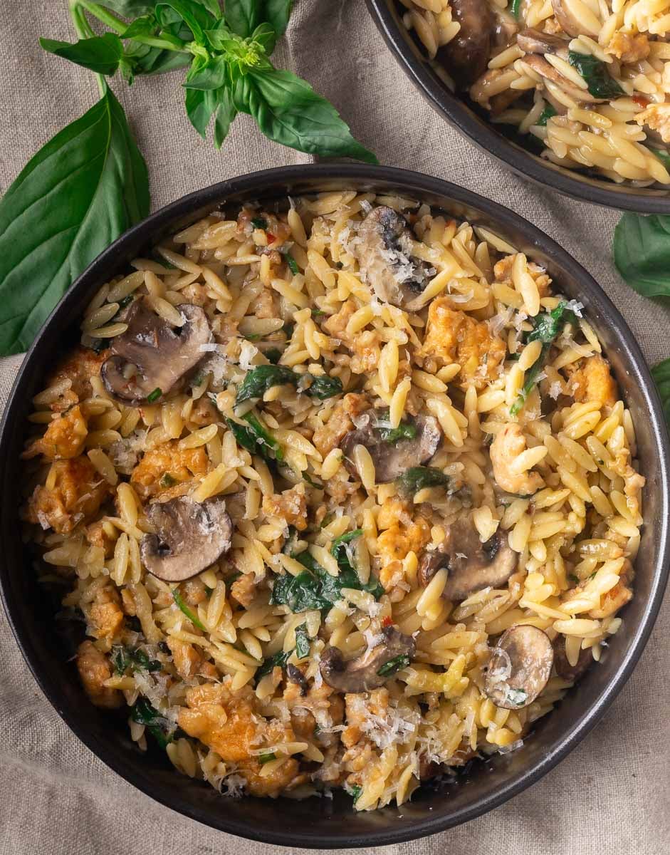 Bowl of chicken sausage and mushroom orzo pasta with mushrooms, spinach, and herbs on a cloth, garnished with fresh basil leaves.