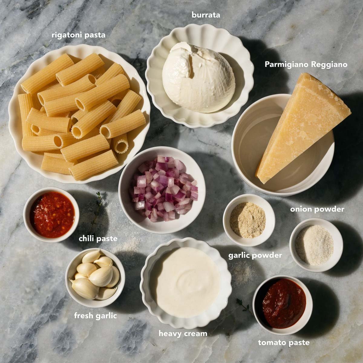 Ingredients for spicy burrata pasta on marble: rigatoni, burrata, Parmigiano, chili paste, diced onion, garlic, cream, tomato paste, powders.