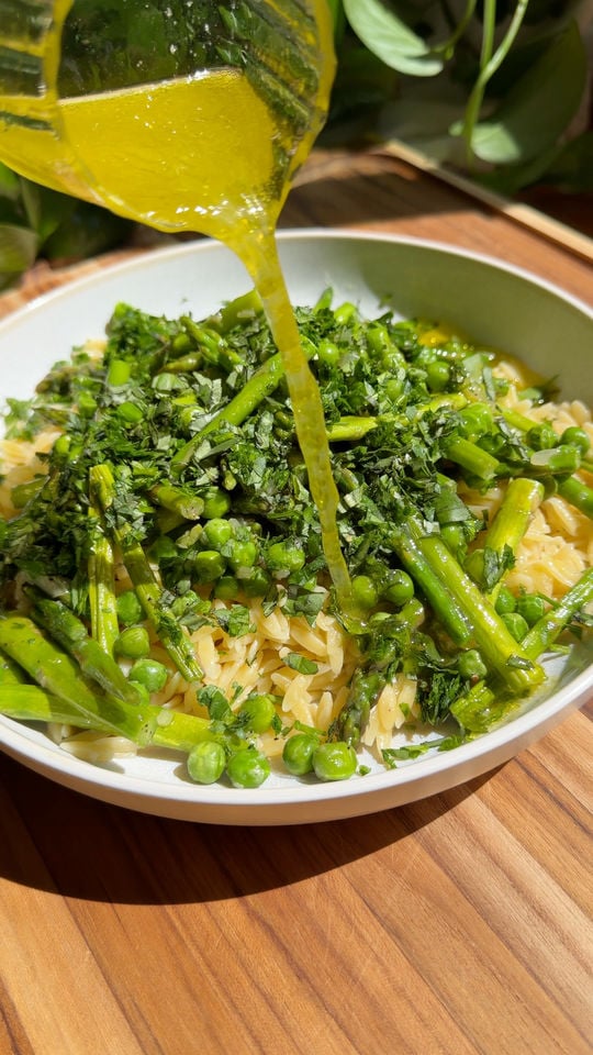 Pouring lemon sauce over pasta with asparagus, peas, and herbs in a white bowl.