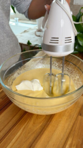 Person mixing batter with handheld mixer in a glass bowl on wooden surface.