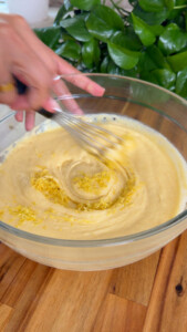 Hand whisking batter with lemon zest in a glass bowl on a wooden surface.