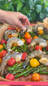 Hand arranging shrimp, cherry tomatoes, and asparagus on a tray with leafy plants in the background.