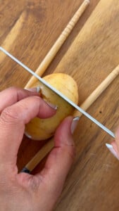 Hand slicing potato on wooden board with knife and chopsticks as guides.