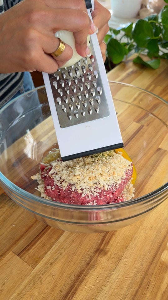 Person grating onion over ground meat and breadcrumbs in a glass bowl on a wooden countertop.