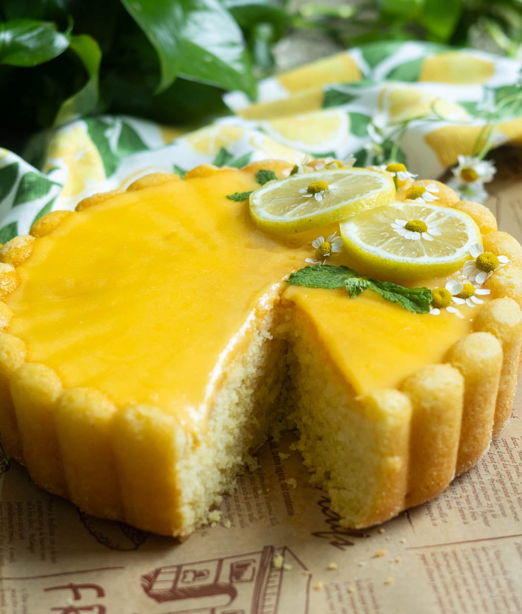 Lemon Semolina Olive Oil Cake with sliced lemon and mint garnish, one slice missing, on a wooden surface with greenery in the background.
