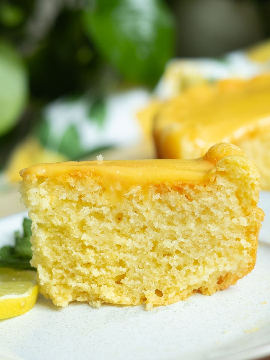 Lemon Semolina Olive Oil Cake with yellow glaze, garnished with lemon slices and mint, on a white plate, green background.