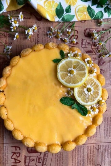 Lemon Semolina Olive Oil Cake with yellow glaze, garnished with lemon slices, mint leaves, chamomile flowers, on wooden table.