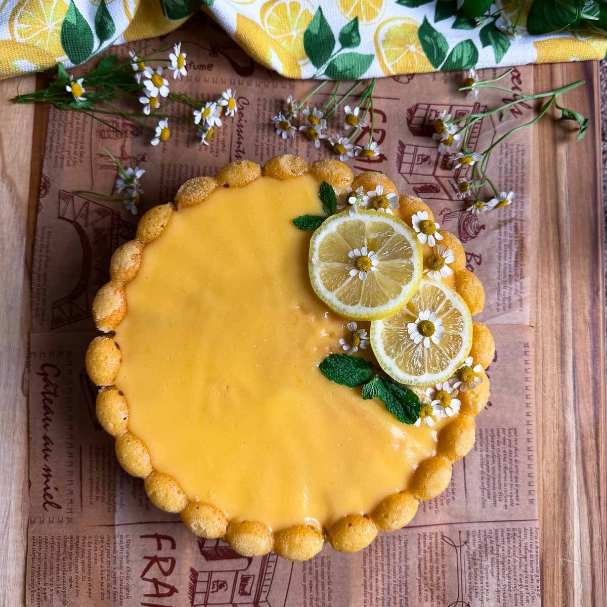 Lemon Semolina Olive Oil Cake with yellow glaze, garnished with lemon slices, mint leaves, chamomile flowers, on wooden table.