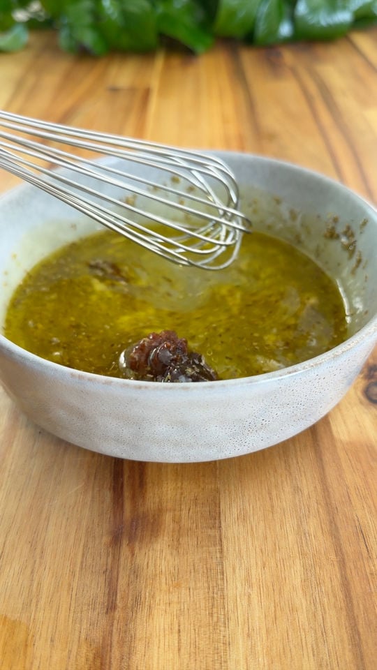 Whisk stirring olive oil and herbs in a ceramic bowl on wooden table.