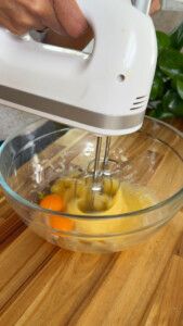 Hand mixer blending eggs and batter in a glass bowl.