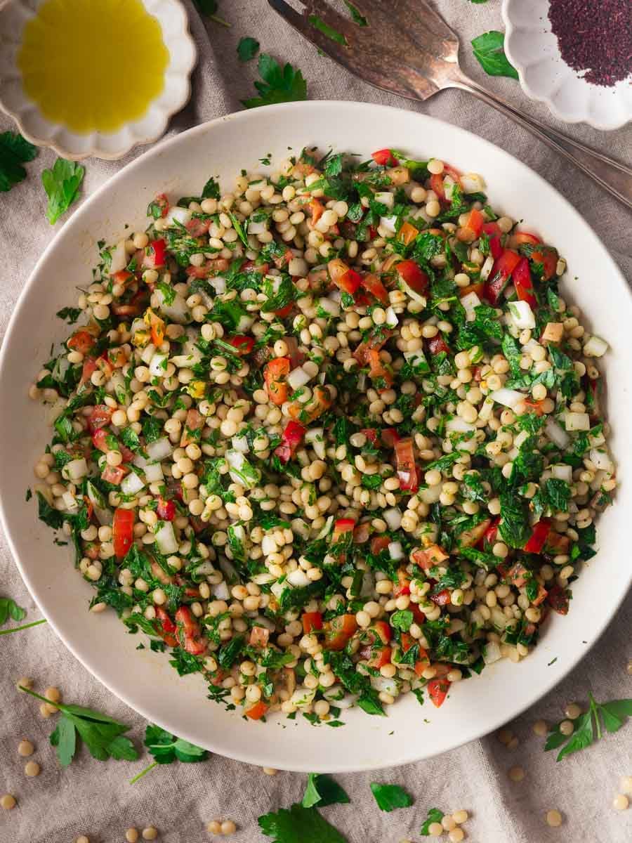Bowl of pearl couscous salad with parsley, tomatoes, onions, and dressing on a linen cloth with herbs and spices nearby.