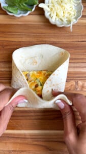 Hands folding a tortilla wrap with egg filling on a wooden board.