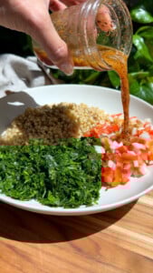 Pouring dressing over couscous, parsley, and diced vegetables in a white bowl.