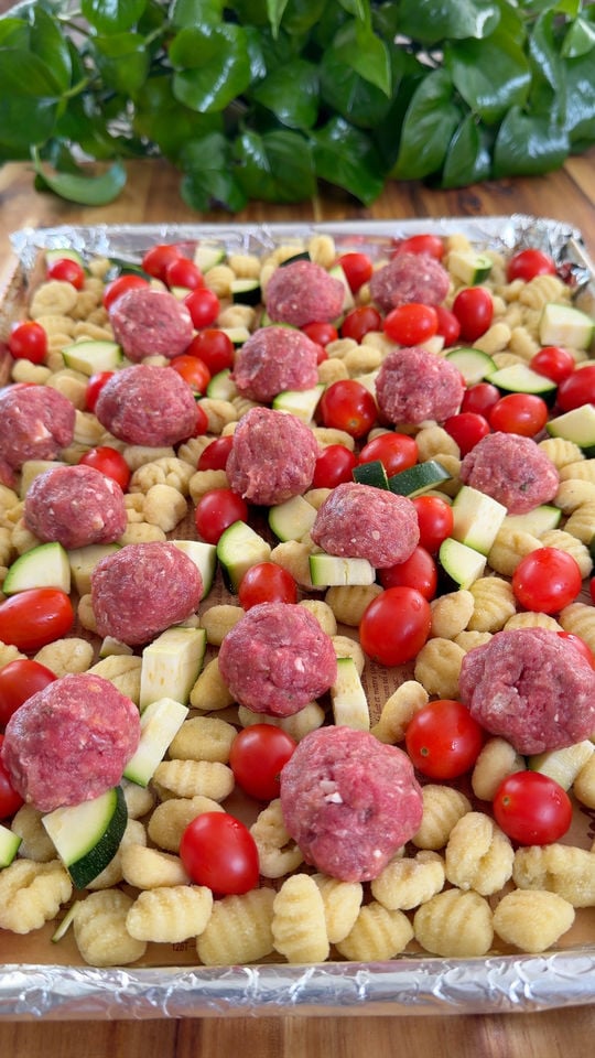 Raw meatballs, gnocchi, cherry tomatoes, zucchini on a foil-lined baking sheet.