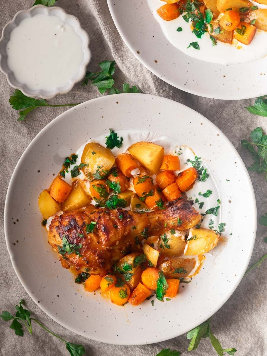Baked red pepper and garlic drumsticks with potatoes, carrots, and parsley on a white plate, side dish of sauce in a small bowl.