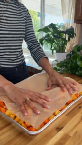 Person covering chopped vegetables and chicken on a baking tray with parchment paper.
