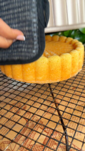 Person removing a sponge cake from a mold using a pot holder on a cooling rack.