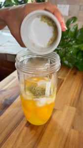 Hand pouring herbs into a blender cup with eggs and Greek yogurt.