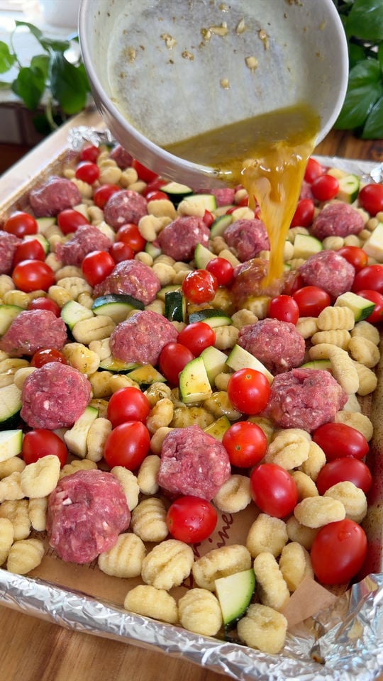 Pouring sauce over a tray of uncooked gnocchi, cherry tomatoes, zucchini, and meatballs.