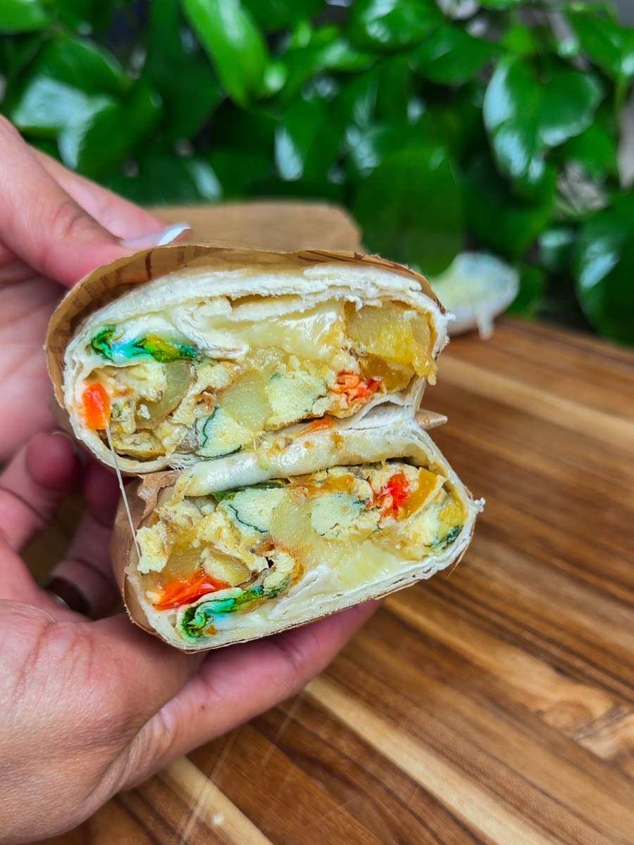 Close-up of a hand holding a sheet pan breakfast burrito with colorful vegetables and cheese on a wooden surface.