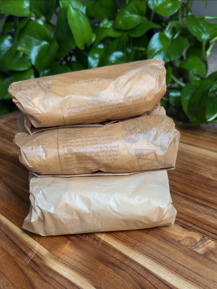 Three brown paper-wrapped sheet pan breakfast burritos stacked on a wooden surface with green leaves in the background.
