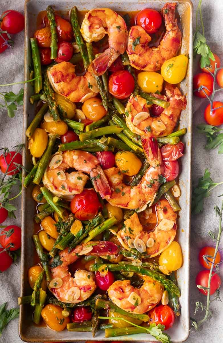 Sheet pan garlic shrimp with asparagus, cherry tomatoes, and sliced garlic garnished with herbs.